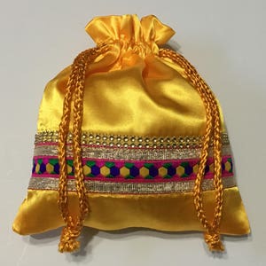 May include: A yellow satin drawstring bag with a colorful embroidered trim. The bag has a gold and pink ribbon and a gold cord.
