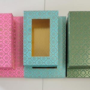 May include: Three decorative boxes in pink, teal, and green, each with a gold filigree pattern. The teal box has a rectangular window. The boxes are arranged side by side, showcasing their contrasting colors and designs.