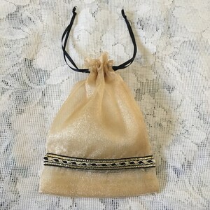 K&ouml;nnte beinhalten: Eine goldene Kordeltasche mit schwarzen Schleifenb&auml;ndern und einem gold-schwarzen Besatz. Die Tasche ist aus einem durchsichtigen Stoff.