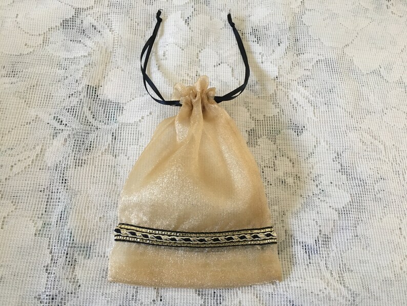 K&ouml;nnte beinhalten: Eine goldene Kordeltasche mit schwarzen Schleifenb&auml;ndern und einem gold-schwarzen Besatz. Die Tasche ist aus einem durchsichtigen Stoff.