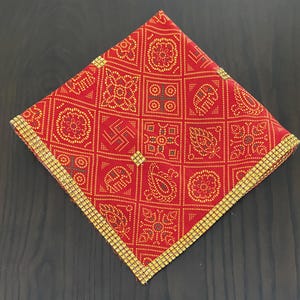 Könnte beinhalten: Ein quadratisches rotes Stofftuch mit einem sich wiederholenden Gittermuster aus gelben geometrischen Formen, Blumenmotiven und Paisley-Mustern. Die Kanten des Tuchs sind mit einer dekorativen Borte aus kleinen, goldfarbenen Perlen versehen.