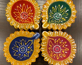May include: Four colorful, hand-painted clay diya lamps. Each lamp features a different color: red, green, blue, and maroon, with a gold rim and a floral design in the center. The lamps are arranged on a dark wooden surface.