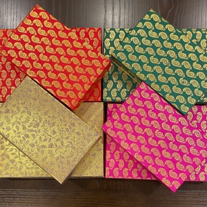 May include: Five decorative boxes with different patterns. The boxes are made of cardboard and have a gold metallic finish. The patterns include paisley, floral, and geometric designs. The boxes are in shades of red, green, pink, and gold.