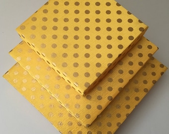 May include: Three yellow square boxes stacked, each with a gold polka dot pattern. The boxes are arranged in a diagonal stack, with the largest box at the bottom and the smallest at the top. The boxes are made of a textured material.