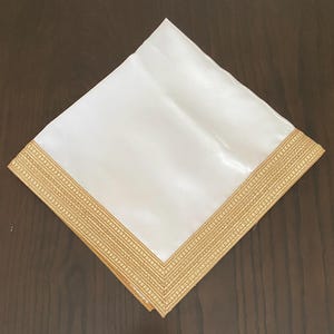 Könnte beinhalten: Eine quadratische weiße Stoffserviette mit einem breiten, dekorativen goldfarbenen Rand mit strukturiertem Webmuster. Die Serviette ist diagonal gefaltet und zeigt den Kontrast zwischen der glatten weißen Mitte und der goldenen Verzierung.