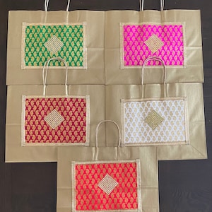 Könnte beinhalten: Satz von fünf goldenen Geschenktüten mit dekorativen, gemusterten Einsätzen. Jede Tasche hat einen Einsatz in einer anderen Farbe mit einem rautenförmigen Design in der Mitte. Die Einsätze sind grün, rosa, rot, weiß und dunkelrot.