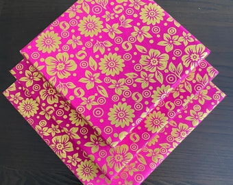 May include: Three folded pink floral patterned napkins. The napkins have a gold floral design. The napkins are stacked on top of each other, with the top napkin angled to show the design. The background is a dark wood surface.