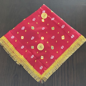 Könnte beinhalten: Ein quadratisches rotes Stofftuch mit einem wiederkehrenden Muster aus kleinen weißen und gelben geometrischen Formen. Das Tuch ist mit zwei runden, goldfarbenen Zier-Medaillons versehen und an allen vier Kanten mit einer goldfarbenen Franse besetzt.