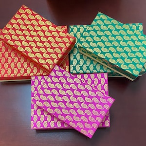 May include: An overhead shot of several decorative boxes in various colors. The boxes are red, green, and pink, each adorned with a gold paisley pattern. The boxes are arranged on a dark brown surface.