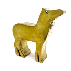 May include: A stylized brass animal figurine with a smooth, polished golden-yellow finish. The figure features a distinct, elongated neck and head, with a single inlaid blue and white eye detail on the side. The body stands on four legs.
