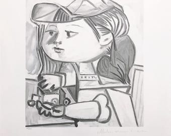 May include: Monochromatic print of a child in a hat, holding a small object. The artwork, in shades of gray, features a stylized portrait with bold lines and shapes. The print has handwritten text in the upper right corner and a signature at the bottom.