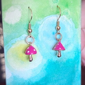 May include: A pair of mushroom-shaped earrings. The earrings feature a pink cap with white polka dots and a gold-colored stem. The earrings are suspended from gold-colored hooks.