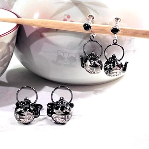 May include: Silver-colored teapot earrings and matching stud earrings. The earrings are shaped like miniature teapots with a handle and spout. The earrings are displayed on a wooden chopstick, with a white bowl in the background.