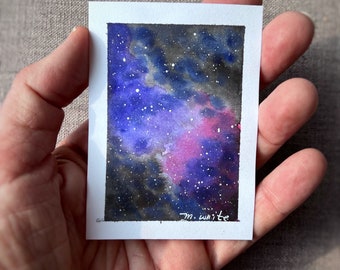 May include: A small watercolor painting of a nebula, featuring shades of purple, blue, and pink with white speckles representing stars. The artwork is on a white rectangular card, held in a hand. The artist's signature, "m. white", is visible.