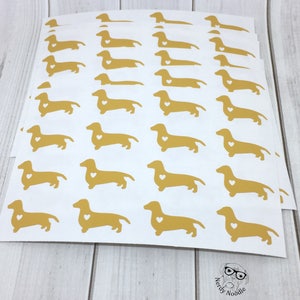 K&ouml;nnte beinhalten: Ein Blatt wei&szlig;es Papier mit mehreren goldenen Dackel-Aufklebern. Jeder Dackel hat ein kleines wei&szlig;es Herz auf der Brust.