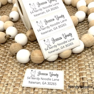 May include: A set of white and brown round wooden beads with a roll of white address labels featuring a black and white illustration of a chicken and the text "Jessica Young, 54 Nerdy Noodle Lane, Newnan, GA 30265".