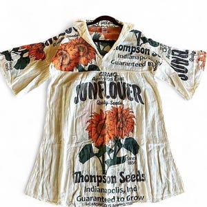 Puede incluir: Vestido túnica color crema con un gran gráfico de girasol en naranja y verde. El vestido tiene mangas anchas y cuello en V. El texto en el vestido incluye "Sunflower" y "Thompson Seeds, Indianapolis, Ind."