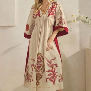 May include: A cream-colored, long dress with a V-neck and elbow-length sleeves. The dress features red and gold embroidered floral patterns on the bodice, sleeves, and skirt. The dress has a flowing silhouette and is paired with white boots.