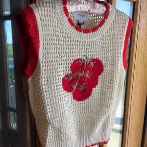 K&ouml;nnte beinhalten: Eine cremefarbene, &auml;rmellose Strickweste mit offener Webstruktur, Rundhalsausschnitt und leuchtend roten Rippb&uuml;ndchen. Auf der Vorderseite befindet sich eine rote, gestickte Tomatengrafik mit gr&uuml;nen Stielen und Bl&auml;ttern. Das Etikett zeigt OTIOR.
