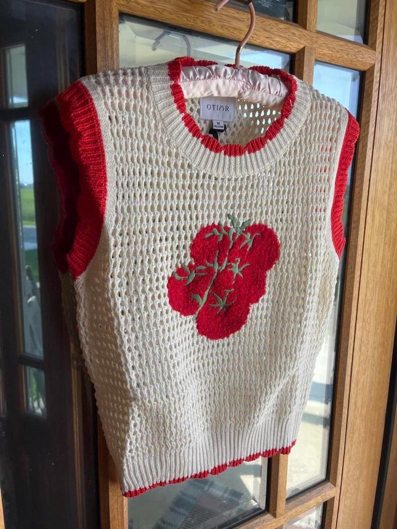 K&ouml;nnte beinhalten: Eine cremefarbene, &auml;rmellose Strickweste mit offener Webstruktur, Rundhalsausschnitt und leuchtend roten Rippb&uuml;ndchen. Auf der Vorderseite befindet sich eine rote, gestickte Tomatengrafik mit gr&uuml;nen Stielen und Bl&auml;ttern. Das Etikett zeigt OTIOR.