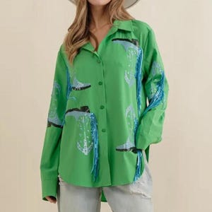 Puede incluir: Una camisa verde brillante con botones, mangas largas y cuello. La camisa presenta un estampado de diseños de botas azules y blancas y flecos azules. Se combina con vaqueros de lavado claro y un sombrero blanco.