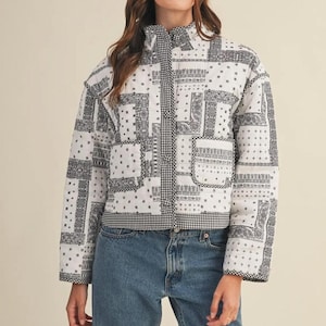 May include: A quilted jacket in a black and white bandana print. The jacket has a high collar, a black and white gingham trim, and two front pockets. The model is wearing blue jeans and a black and white baseball cap.