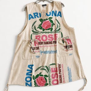 Puede incluir: Delantal beige con texto azul y rosa, con la palabra "ARIZONA" y una gran rosa rosa. El delantal tiene dos bolsillos y está colgado de una percha de metal.