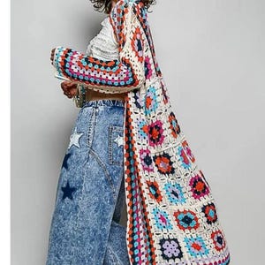 May include: A long, open-front cardigan made of multi-colored crochet granny squares in shades of red, blue, pink, and orange with cream borders. The cardigan is paired with a white cropped top and acid-wash blue jeans featuring dark blue star patches.
