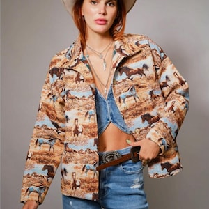 May include: A long-sleeved jacket featuring an all-over print of horses in a desert landscape with shades of brown, tan, and light blue. The jacket has a collared neckline, a button-front closure, and a structured fit with a repeating pattern of horses.