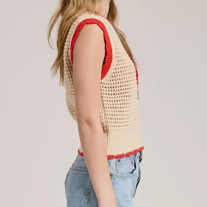 K&ouml;nnte beinhalten: Eine &auml;rmellose H&auml;kelweste in hellem Beige mit einer ausgepr&auml;gten Netzstruktur. Das Kleidungsst&uuml;ck ist mit einem leuchtend roten Rippstricksaum an den Armausschnitten, dem Halsausschnitt und dem unteren Saum versehen, kombiniert mit hellblauen Jeansshorts.