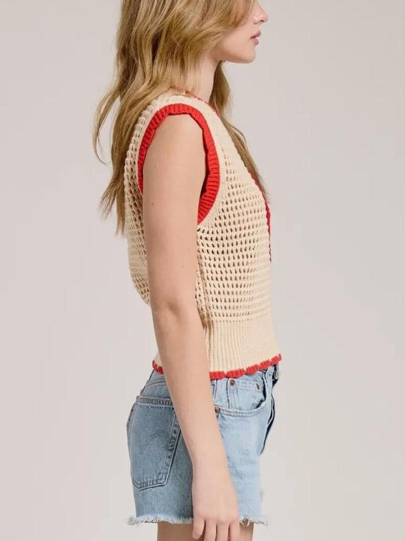 K&ouml;nnte beinhalten: Eine &auml;rmellose H&auml;kelweste in hellem Beige mit einer ausgepr&auml;gten Netzstruktur. Das Kleidungsst&uuml;ck ist mit einem leuchtend roten Rippstricksaum an den Armausschnitten, dem Halsausschnitt und dem unteren Saum versehen, kombiniert mit hellblauen Jeansshorts.