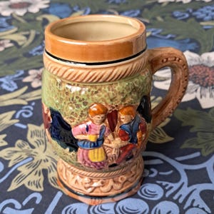 May include: A ceramic beer stein with a handle. The stein features a detailed relief scene of figures in traditional clothing, with brown, green, and tan colours. The top and base have decorative trim.