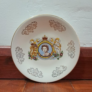 May include: A white commemorative plate with a gold rim and decorative gold filigree patterns. The center features a portrait of Queen Elizabeth II within a crest, with text that reads "To Commemorate The Silver Jubilee of Queen Elizabeth II 1977."