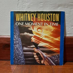 May include: A vinyl record cover featuring a hand holding a torch with flames. The cover is blue and white with the text "Whitney Houston" and "One Moment in Time" in bold, yellow letters.