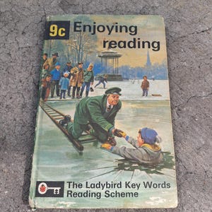 Puede incluir: Portada de libro vintage "Enjoying Reading". La ilustración muestra una escena invernal con gente en un lago helado y un hombre ayudando a un niño. El libro es parte de "The Ladybird Key Words Reading Scheme".