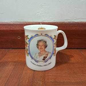 Puede incluir: Taza de cerámica blanca con un retrato de la reina Isabel II dentro de un marco azul y dorado, con el texto "Golden Jubilee 1952-2002". La taza tiene un asa blanca y está decorada con motivos florales.