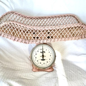 Op de afbeelding: Een roze rieten mandje staat op een vintage roze en witte babyweegschaal. De weegschaal heeft een rond gezicht met zwarte cijfers en een zilveren naald.