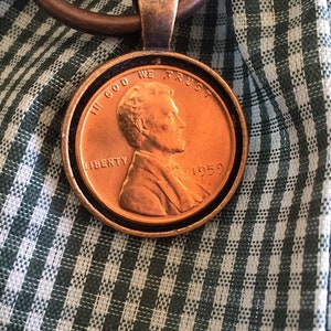 May include: A copper-colored 1959 penny pendant with the words "In God We Trust" and "Liberty" on the front. The pendant is attached to a bronze-colored ring.