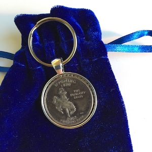 May include: A silver key chain with a blue velvet pouch. The key chain features a silver coin with the image of a cowboy riding a bucking bronco. The coin reads "Wyoming 1890 The Equality State 2007 E Pluribus Unum".