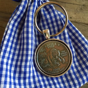 Op de afbeelding: Een zilveren sleutelhanger met een 25 cent munt uit Alaska van 2008 met een bruine beer en de tekst "Alaska 1959 The Great Land" op een blauwe en witte geruite stoffen tas.