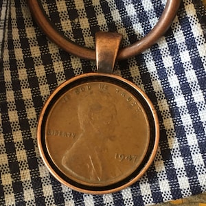 May include: A copper-toned pendant featuring a 1947 US penny with the inscription "In God We Trust" and "Liberty".