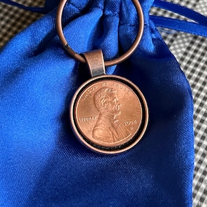 Könnte beinhalten: Ein Schlüsselanhänger aus Kupfer mit einem blauen Samtbeutel. Der Penny ist aus dem Jahr 2004 und trägt die Aufschrift "In God We Trust" und "Liberty".