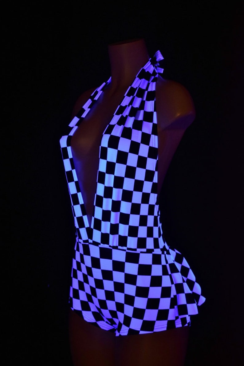 May include: A halter-style romper featuring a black and white checkered pattern. The garment has a deep V-neckline, a tie closure at the back of the neck, and a ruffled hemline. The fabric appears to be illuminated under ultraviolet light.
