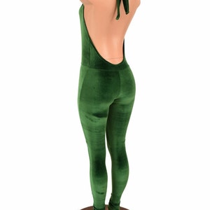 May include: A green velvet jumpsuit with a halter neck and open back. The jumpsuit has a form-fitting design and a bow detail at the neck. The mannequin is standing on a wooden base.