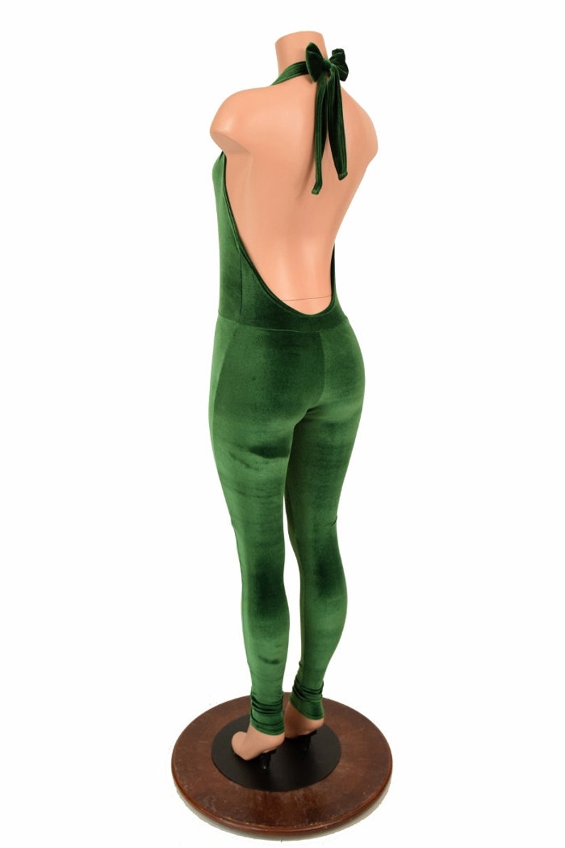 May include: A green velvet jumpsuit with a halter neck and open back. The jumpsuit has a form-fitting design and a bow detail at the neck. The mannequin is standing on a wooden base.