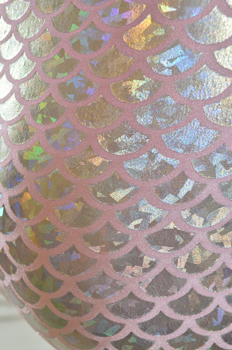 Pu&ograve; includere: Primo piano di un tessuto rosa con un motivo a squame di sirena olografico. Le squame sono iridescenti e brillano alla luce.