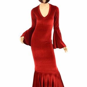 Op de afbeelding: Een lange, rode fluwelen jurk met een V-hals en uitlopende mouwen. De jurk heeft een rok in zeemeermin-stijl en wordt gepresenteerd op een mannequin. De jurk heeft een diepe rode tint.