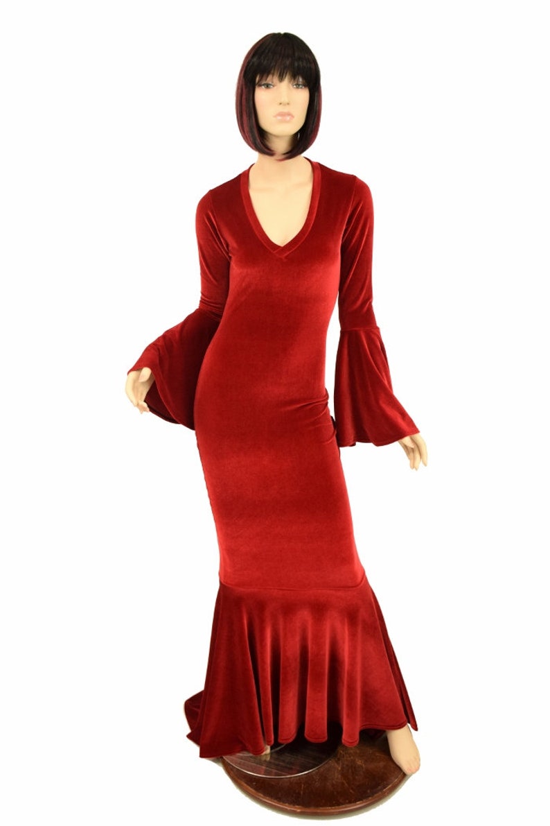 Op de afbeelding: Een lange, rode fluwelen jurk met een V-hals en uitlopende mouwen. De jurk heeft een rok in zeemeermin-stijl en wordt gepresenteerd op een mannequin. De jurk heeft een diepe rode tint.