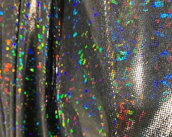 May include: Close-up of a shimmering silver fabric with a holographic effect. The fabric is covered in small, colorful, iridescent dots that reflect light, creating a dazzling display. The material appears to be draped, with folds and shadows.