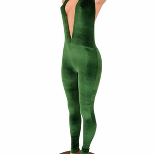 May include: A green velvet jumpsuit with a plunging neckline and halter top design. The one-piece garment features a form-fitting silhouette and long legs. The jumpsuit is displayed on a mannequin with a wooden base.